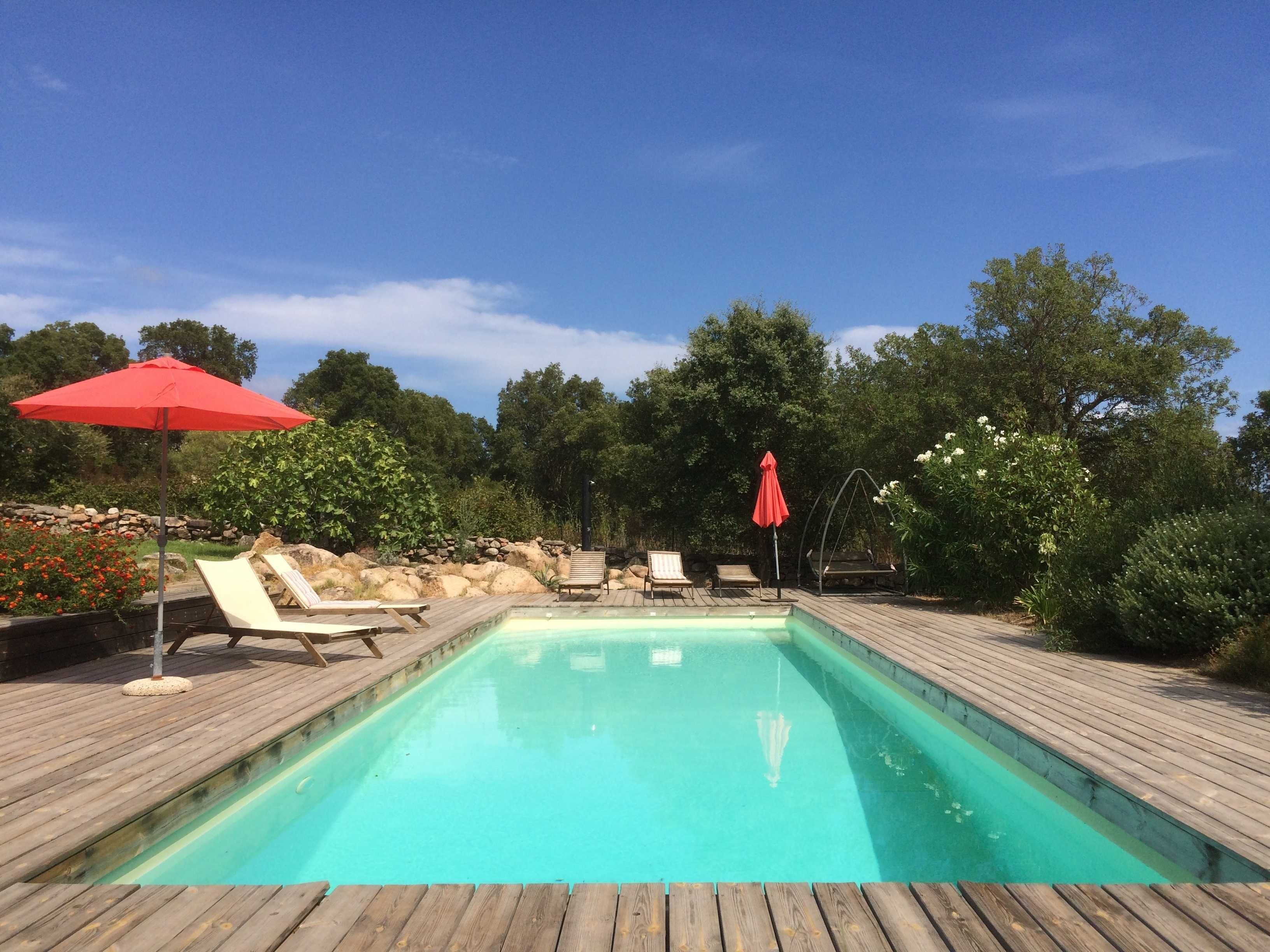 Una piscina rectangular con una terraza de madera alrededor, dos sombrillas rojas y varias sillas de descanso.