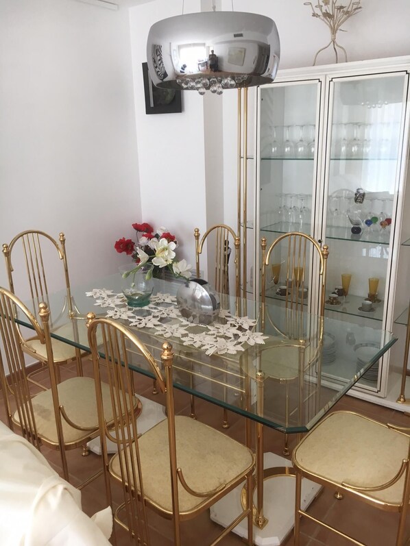 A dining area with a glass table surrounded by gold chairs, a glass cabinet with glassware, and a decorative mirror above the table.