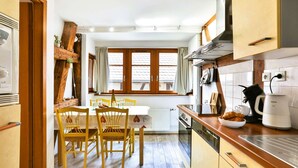 A compact kitchen with wooden cabinets, a dining table with chairs, a window with curtains, and a coffee machine.