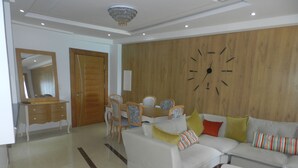 A modern living room with a sofa, a dining table with chairs, a wooden wall clock, and a mirror.