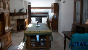 Un comedor con una mesa larga, sillas y una chimenea. Se observan armarios de madera y un techo con vigas a la vista.