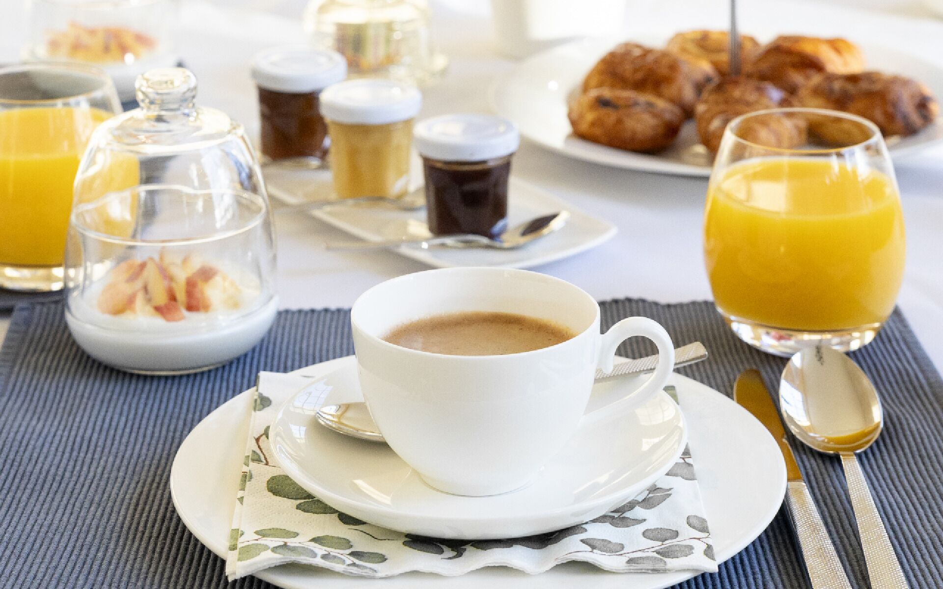 Une table dressée pour le petit-déjeuner, avec une tasse de café, un verre de jus d’orange, des croissants et un verre de lait.