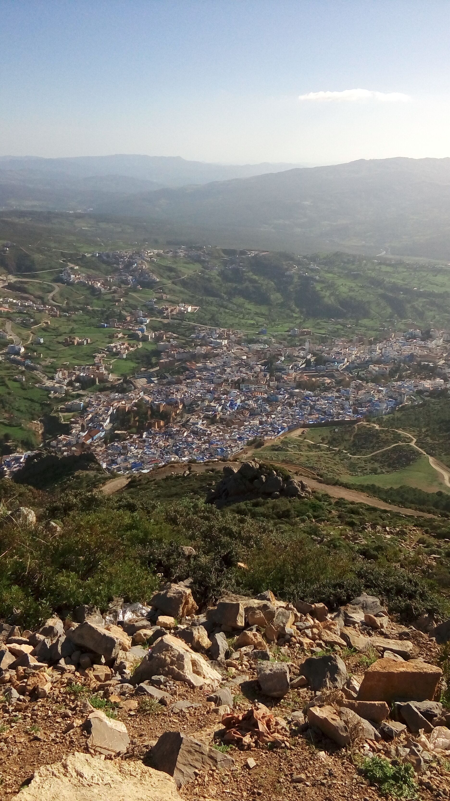 A panoramic view of a town with a mix of residential and commercial buildings, surrounded by green hills and a clear sky.