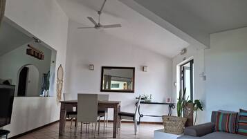 A spacious living area with a dining table, chairs, a sofa, a ceiling fan, and a large mirror on the wall.