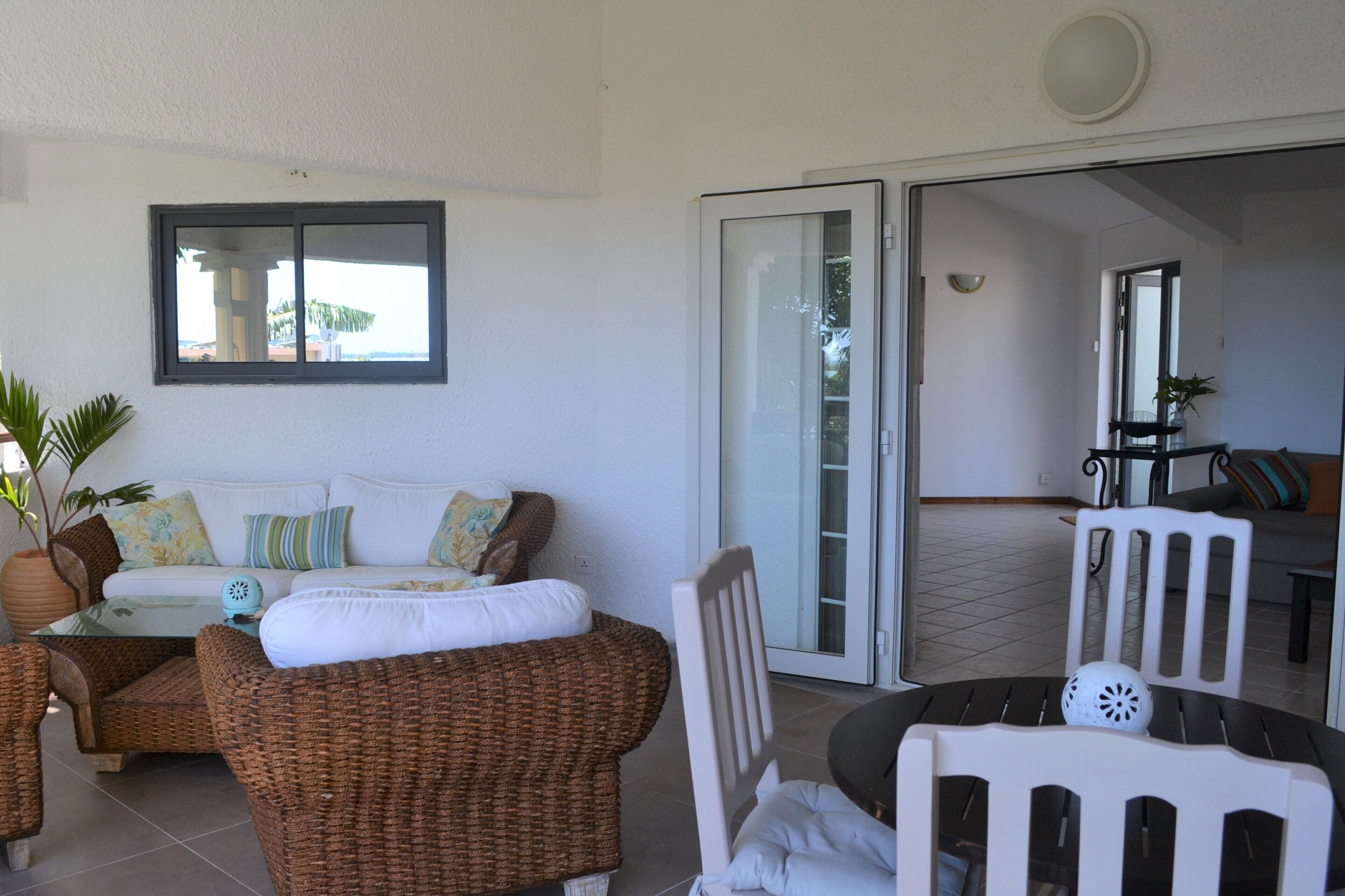 A room with a white sofa, wicker chairs, a glass table, and a sliding glass door leading to a balcony.
