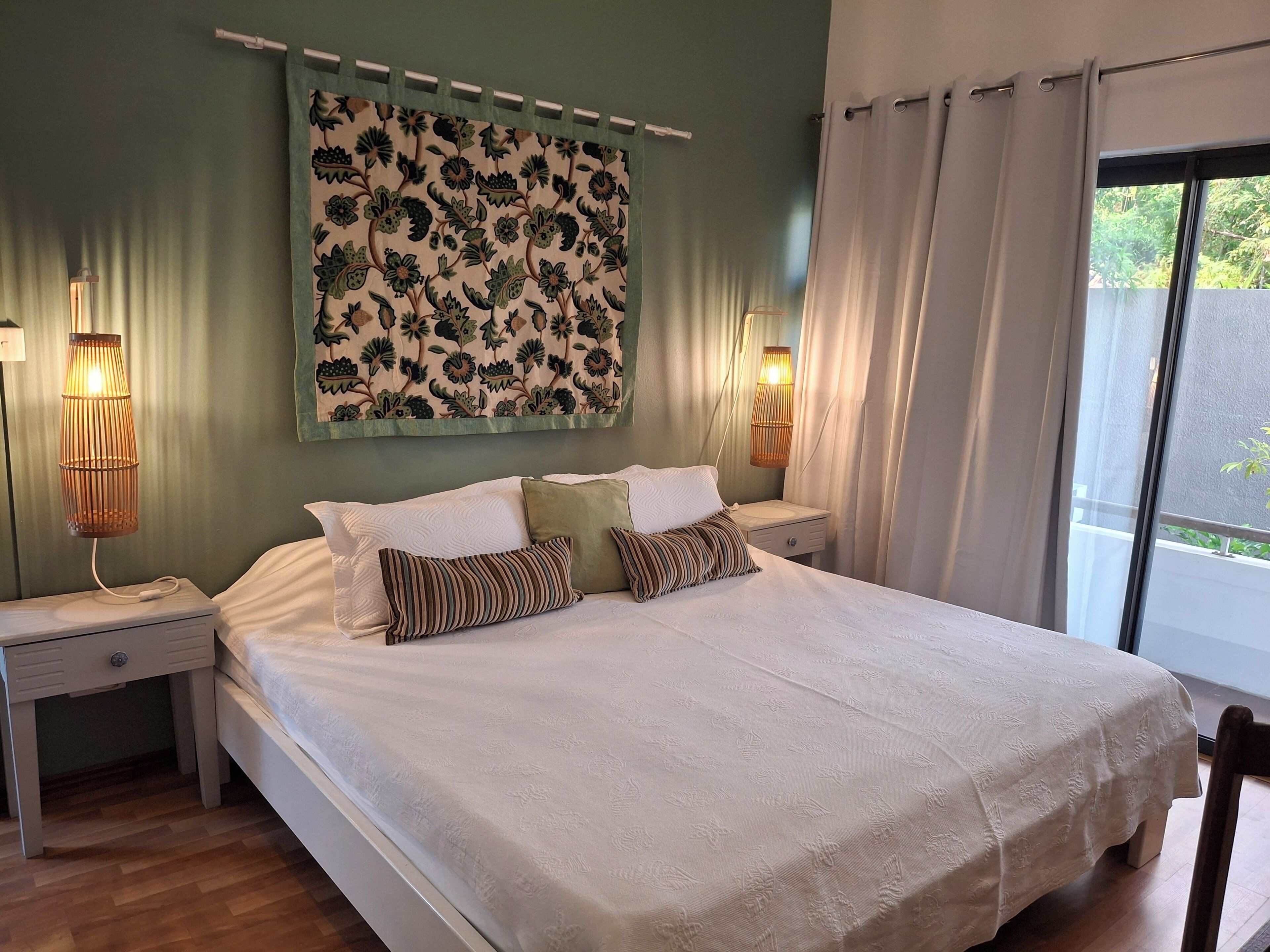 Une chambre à coucher avec un grand lit, deux tables de chevet, une lampe fixée au mur et une fenêtre avec des rideaux.