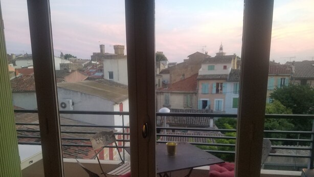 Un balcon avec une table et des chaises, donnant sur un paysage urbain avec des bâtiments et un ciel dégagé.