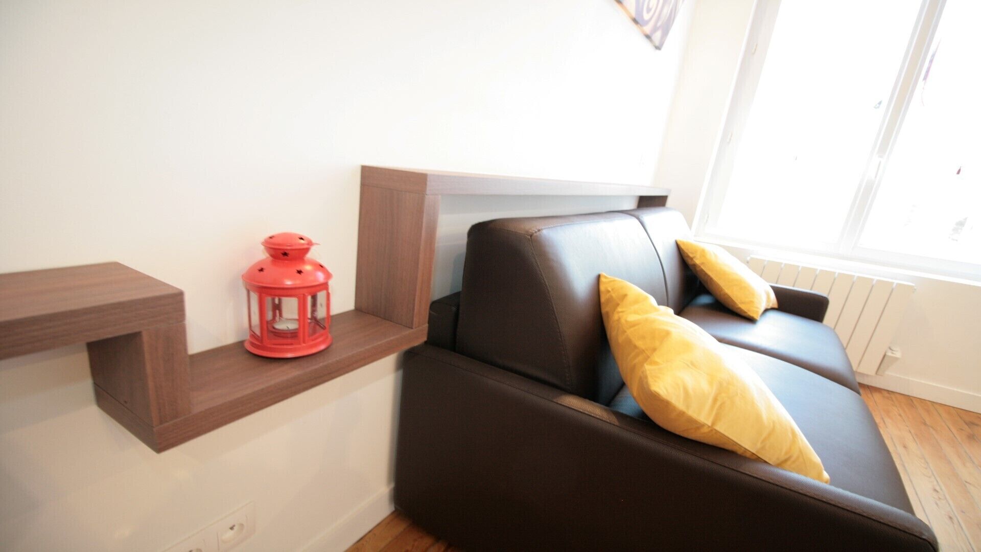 Un salon moderne avec un canapé en cuir marron, deux coussins jaunes, une lanterne rouge et une étagère en bois.