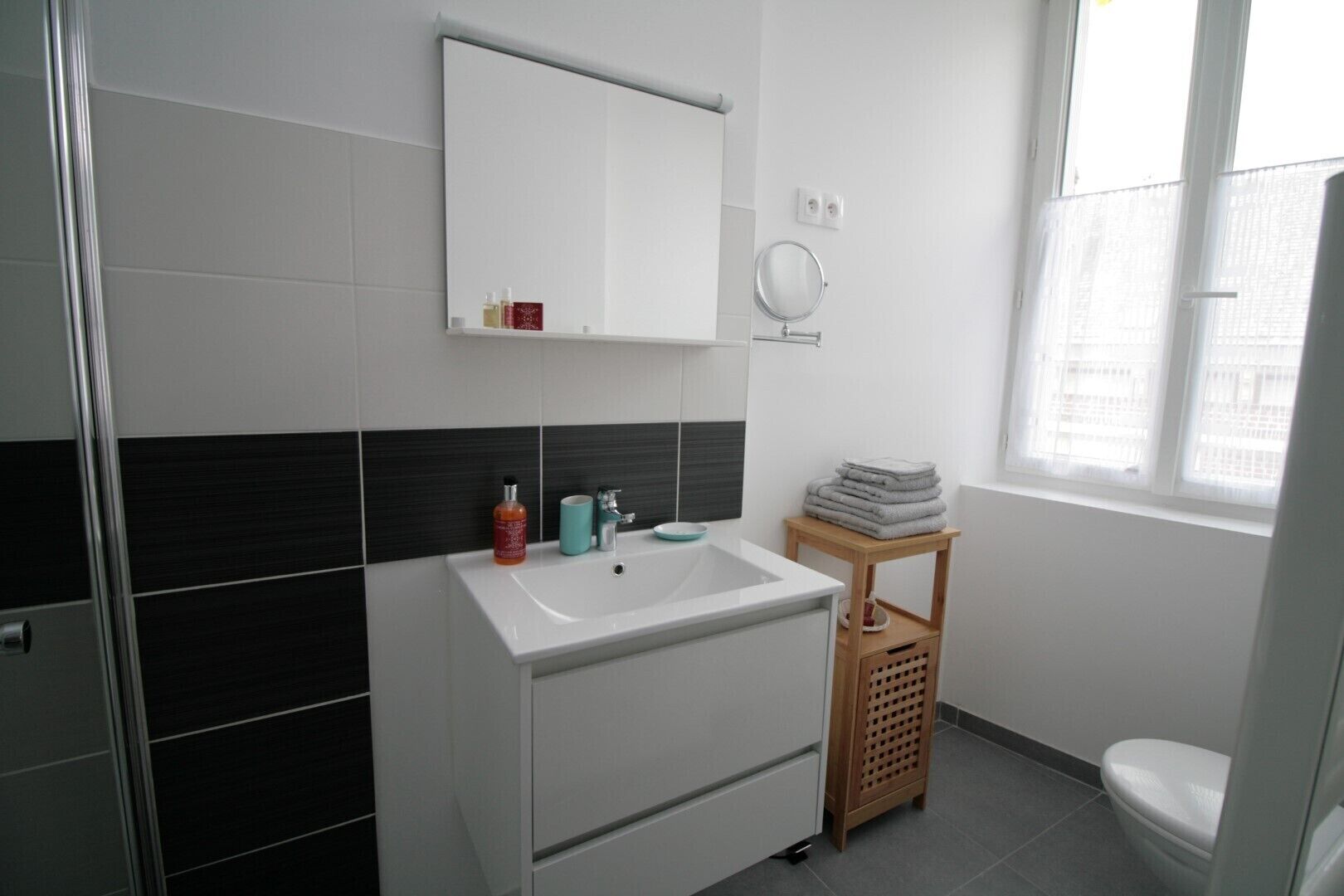 Ein Badezimmer mit einem weißen Waschbecken, einem Spiegel darüber, einer kleinen Holzablage mit Handtüchern und einer Toilette.