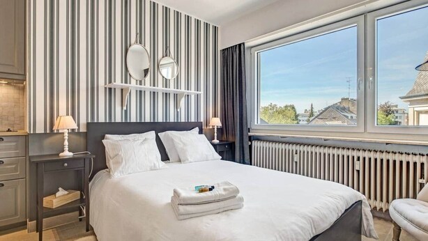 A modern hotel room with a large bed, two bedside tables, a dresser, and a view of the city through a large window. - Studio Chic super central near cathedral (Luxembourg)