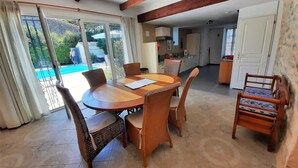 A dining area with a round wooden table and wicker chairs, a kitchen in the background, and a view of a swimming pool through large glass doors.