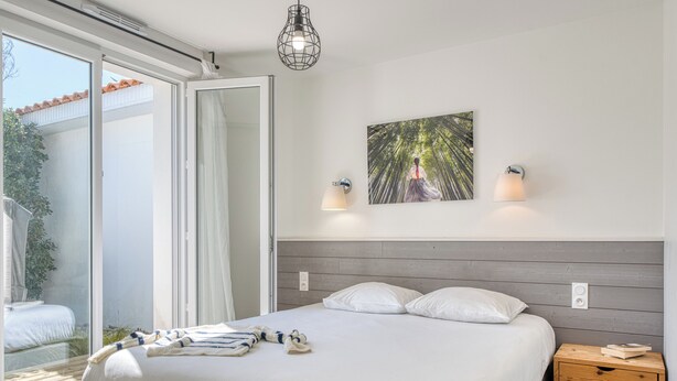 A bedroom with a bed, a wooden nightstand, a wall-mounted light, and a large window.