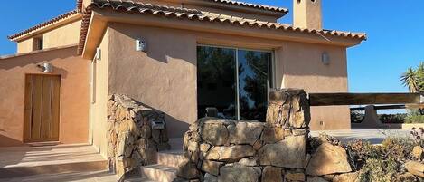 A stucco building with a tiled roof, a stone wall entrance, and a clear sky.