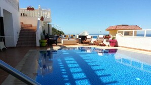 Una piscina con vista despejada al mar, una escalera que lleva a una terraza superior y sillas de descanso a un lado.