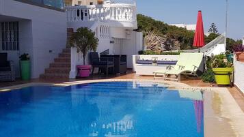 A swimming pool with lounge chairs and a red umbrella, surrounded by a white building with a balcony and steps.