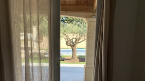 Blick durch ein Fenster mit transparenten Vorhängen, der einen Baum und ein blaues Schwimmbecken im Hintergrund zeigt.
