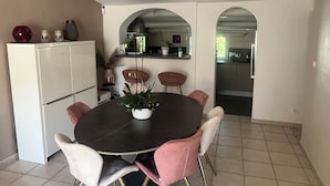 Ein Essbereich mit einem runden Tisch, sechs Stühlen und einer Zimmerpflanze. Ein weißer Kühlschrank befindet sich links, und durch einen Durchgang ist eine Küche erkennbar.