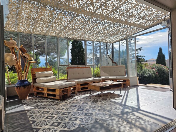 A sunlit room with a glass enclosure, featuring a sofa set, a wooden coffee table, and a decorative vase with wooden utensils.