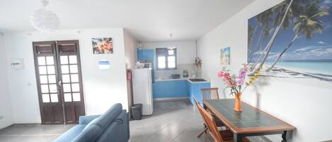 En stue med sofa, spisebord med stole, et køkken i baggrunden og et maleri med et strandmotiv på væggen.