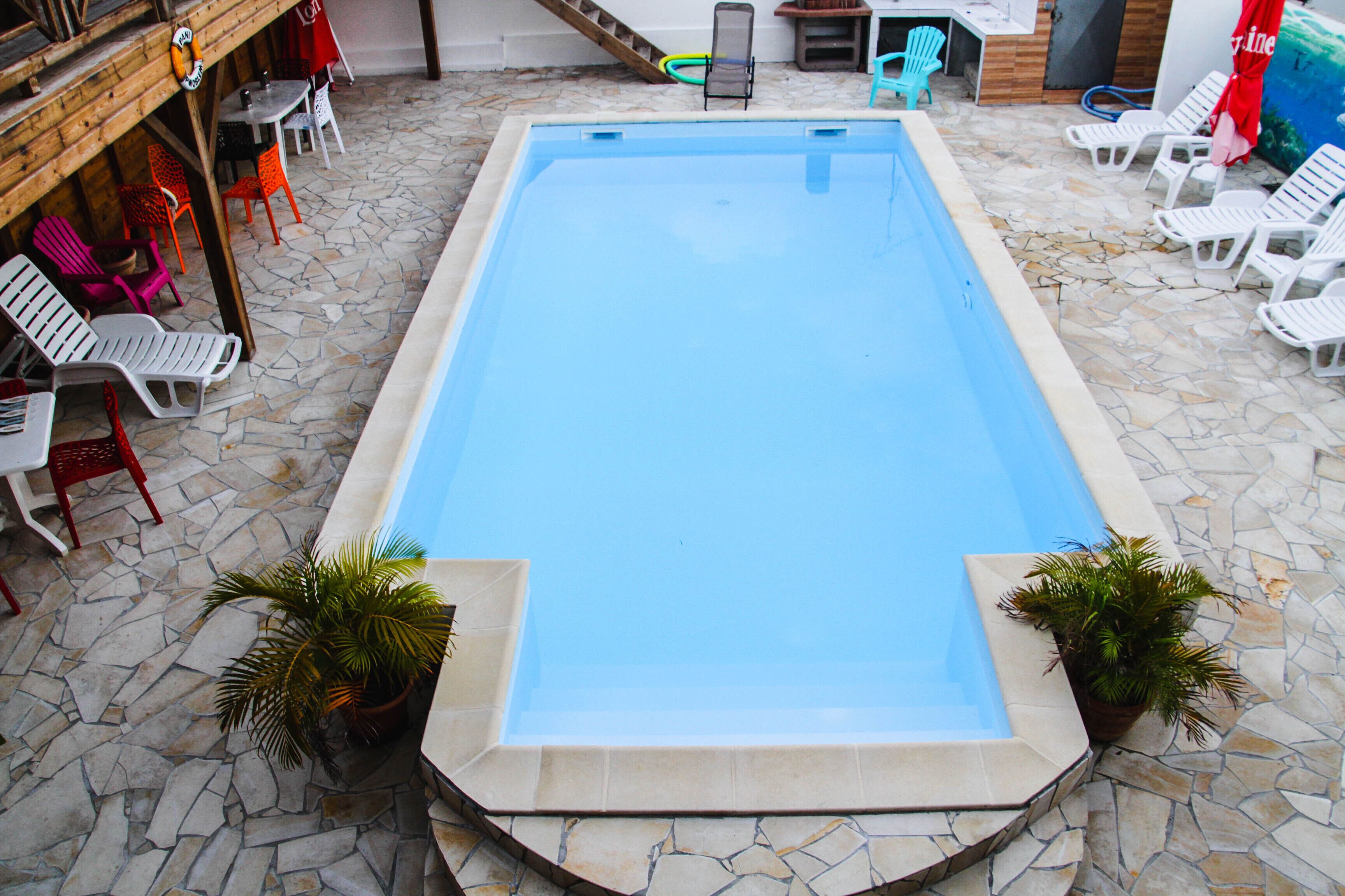A rectangular swimming pool with a stone floor, surrounded by white lounge chairs and potted plants.
