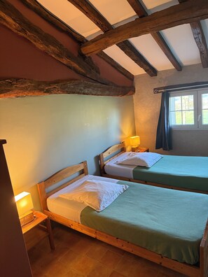 A room with two wooden beds, a small table, and a window with curtains. - Gîte de groupe, 37 places, 13 rooms (La Romieu)
