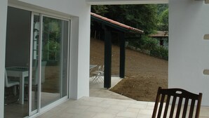 A modern building with a patio area featuring a wooden chair, large glass doors, and a view of a garden.