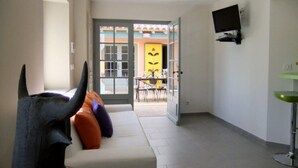 Un salon moderne avec un canapé, un téléviseur fixé au mur et un coin repas visible à travers une porte ouverte.
