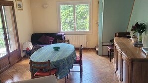 Ruokailutilassa on pyöreä pöytä, jonka päällä on vihreä pöytäliina, ja pöydän ympärillä on puisia tuoleja. Sohva on sijoitettu seinää vasten, ja oikealla puolella näkyy puinen kaappi.