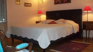 Ein ordentlich gemachtes Bett mit weißer Bettdecke und Kissen, zwei Nachttische mit Lampen, ein gerahmtes Bild an der Wand und ein Sessel mit blauem Sitzkissen.
