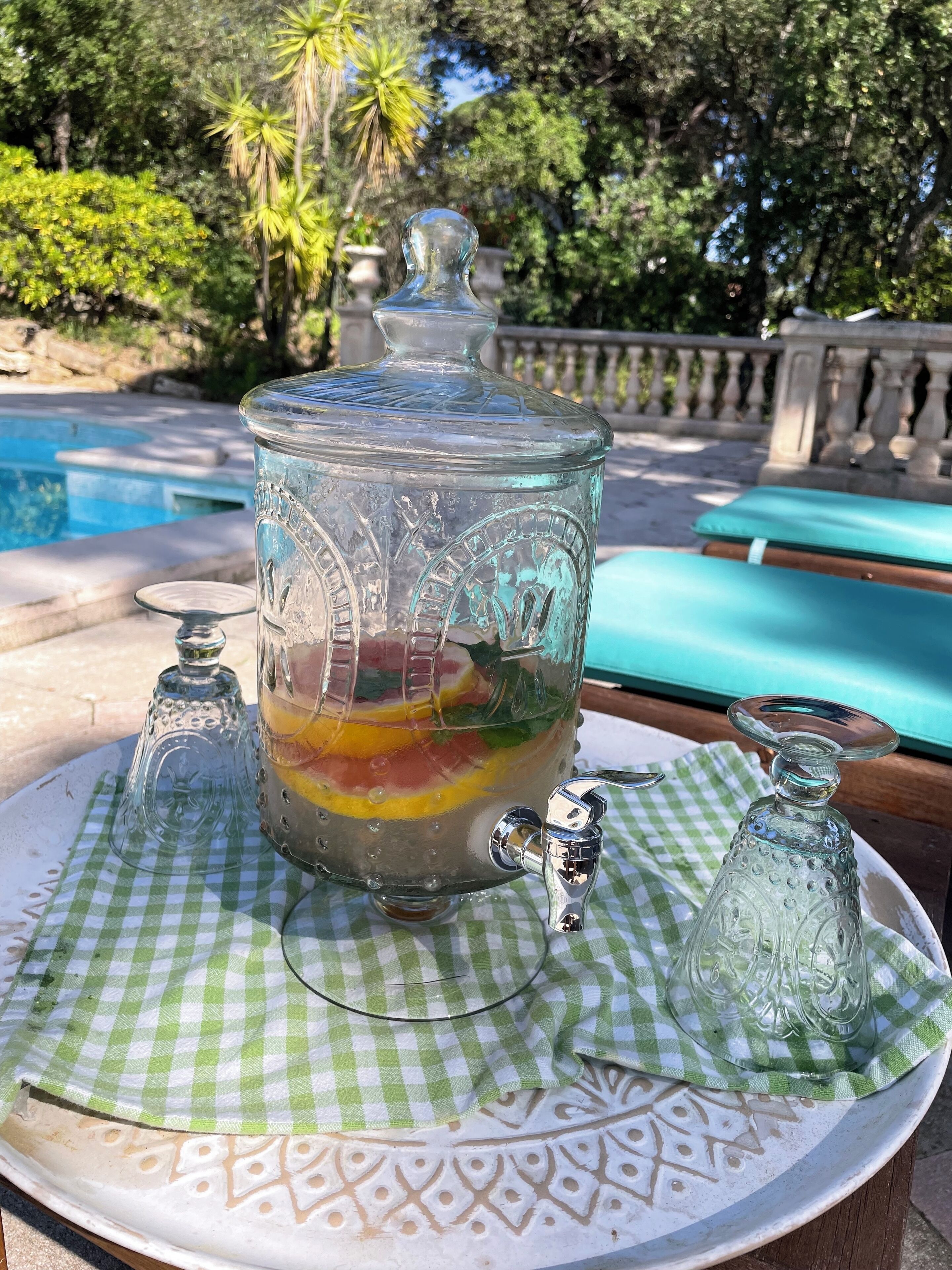 Un bol de ponche de vidrio con capas de fruta sobre un mantel a cuadros, un salero y una cuchara, colocados al aire libre cerca de una piscina.