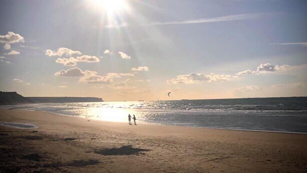 Une plage avec un ciel dégagé, du soleil et quelques personnes qui se promènent.