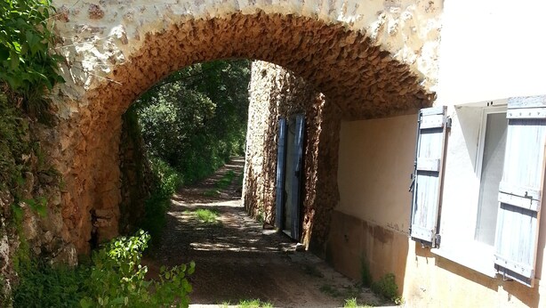 Un arc en pierre rustique qui mène à un sentier étroit, entouré de verdure et bordé d’un bâtiment aux volets en bois.