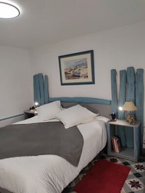 A neatly made bed with pillows and a blanket, a bedside table with a lamp, a framed painting on the wall, and a patterned rug on the floor.