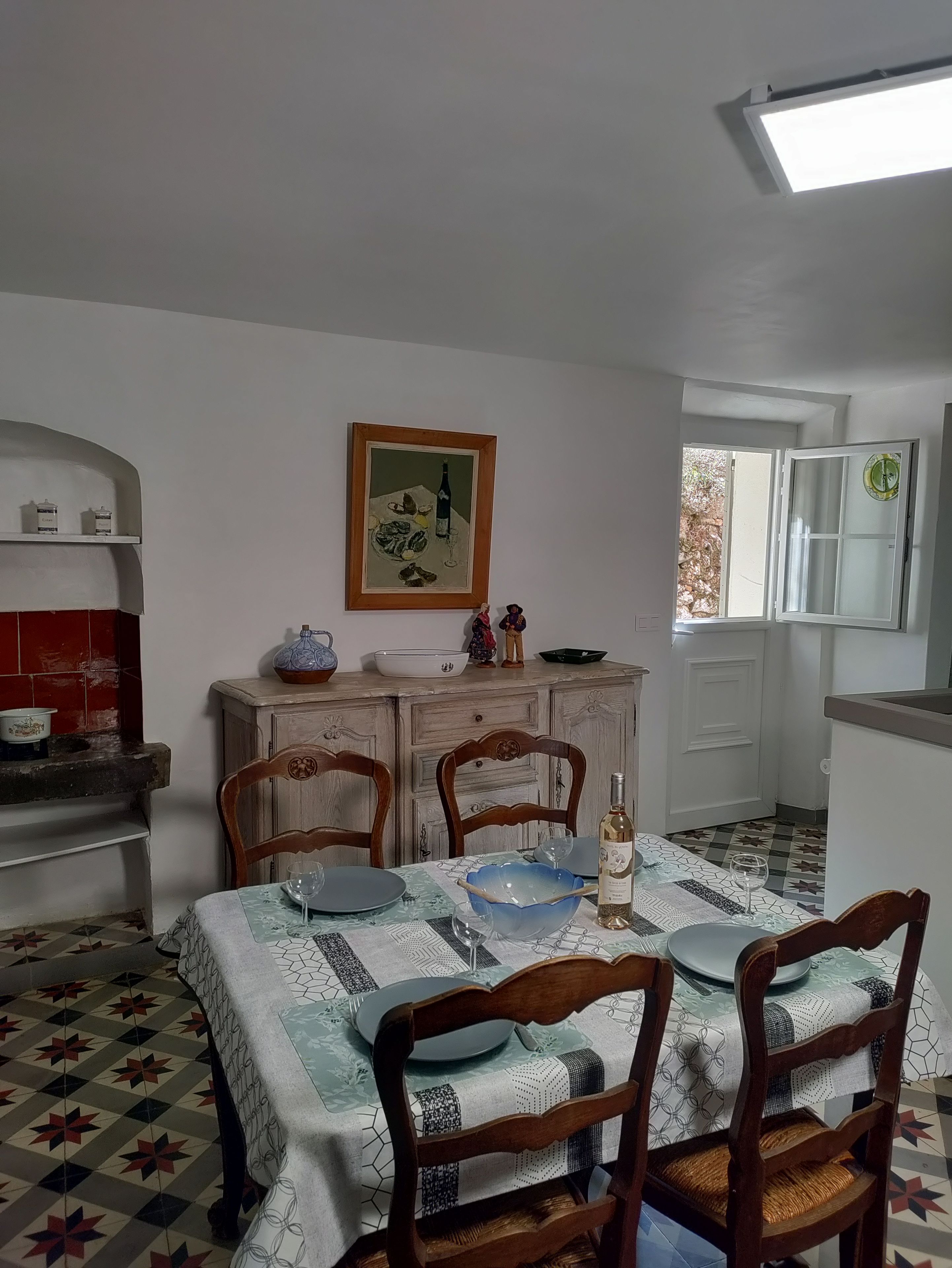 Un comedor con una mesa dispuesta para cuatro, sillas, un mueble con utensilios de cocina, un cuadro en la pared y una ventana con vistas.