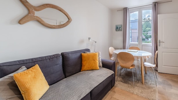 Una sala de estar moderna con un sofá gris, cojines amarillos, un espejo con forma de pez de madera y una zona de comedor con una mesa redonda y sillas.