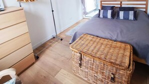 Un dormitorio con cama, cómoda de madera, lámpara de pie, ventana con cortinas e imagen colgada en la pared.
