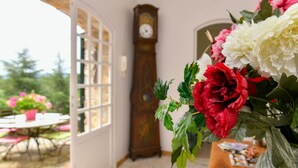 Une pièce avec une grande fenêtre, une horloge en bois et un vase de fleurs.