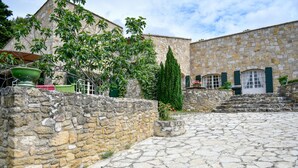 Une cour bordée de murs en pierre, dotée d’un sol en pierre et abritant un bâtiment à la façade en pierre et aux volets verts.