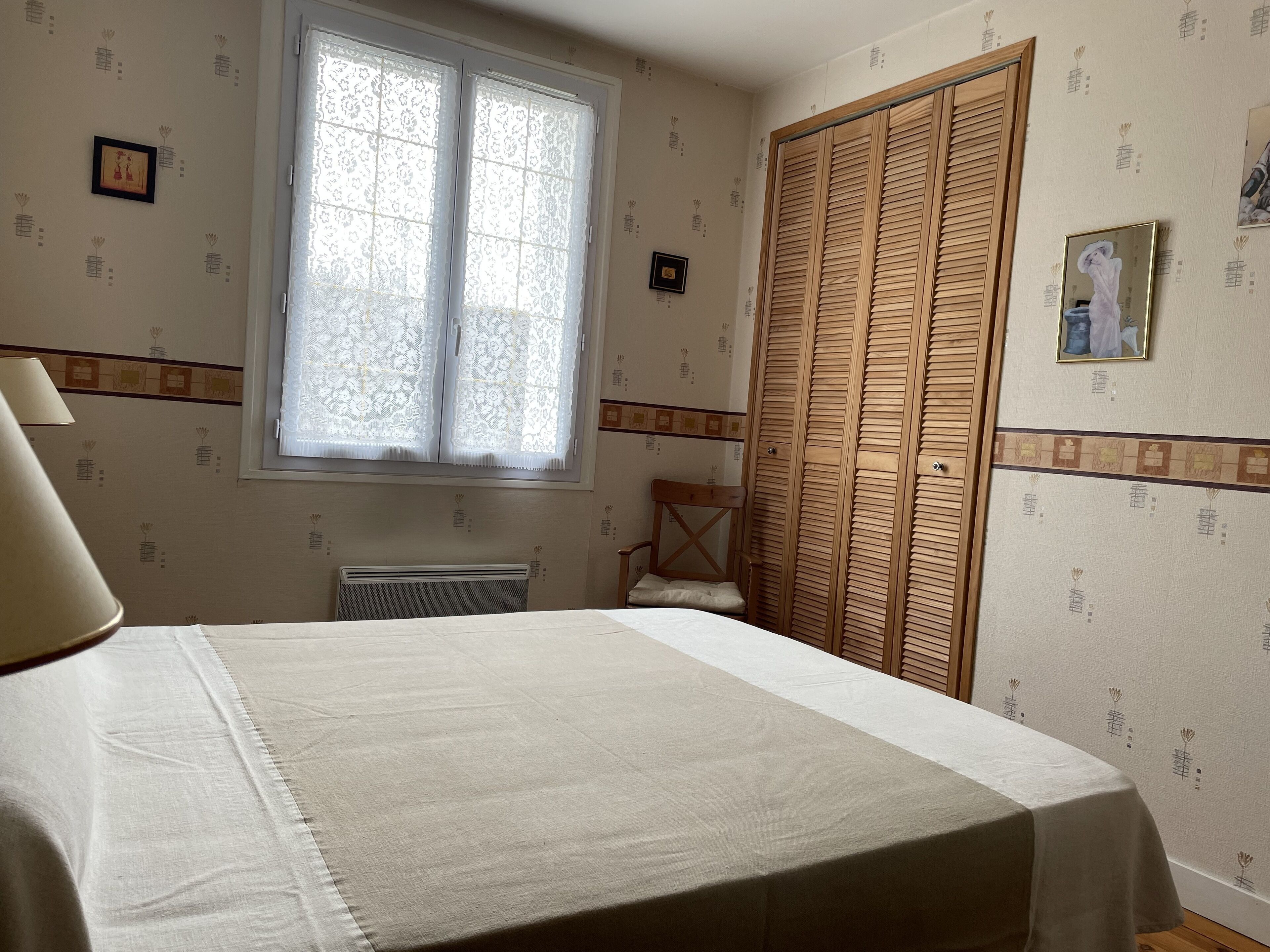 A bedroom with a bed, a window with lace curtains, a radiator, a wooden wardrobe, and patterned wallpaper.