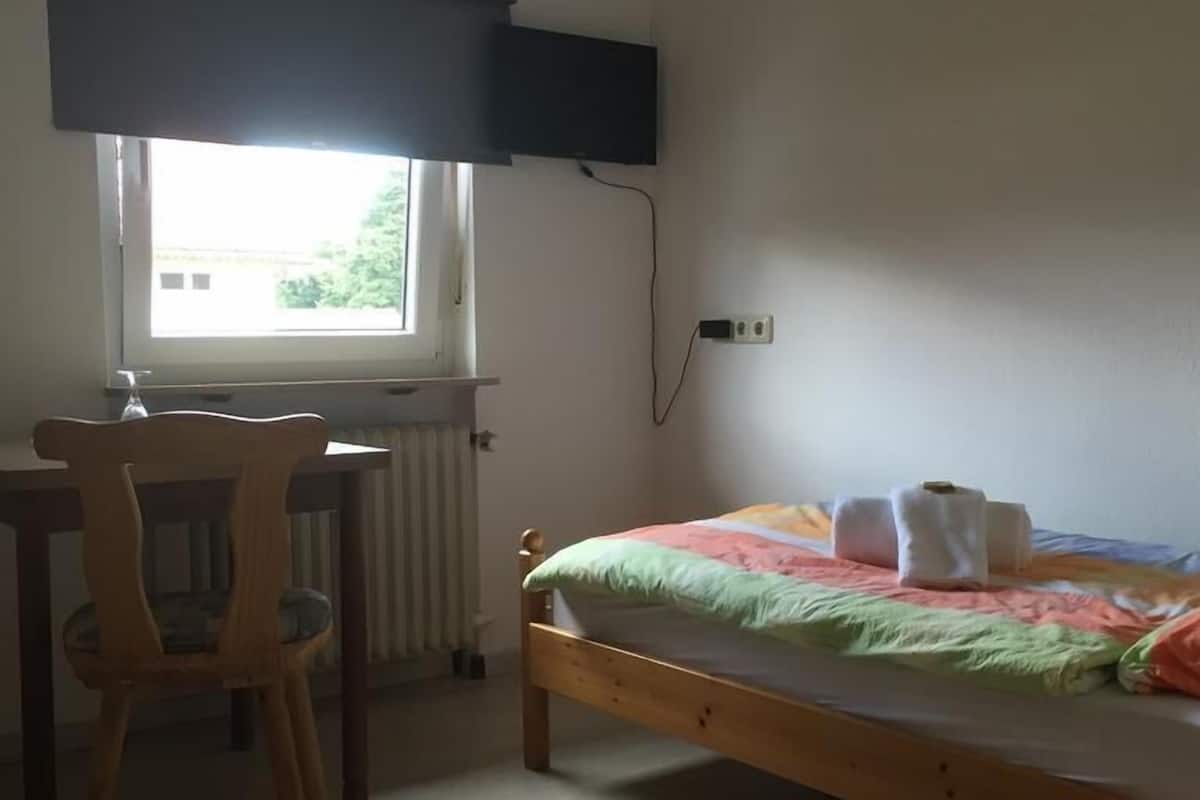 A bedroom with a bed, a chair, a desk, a window with a blind, and a wall-mounted TV.