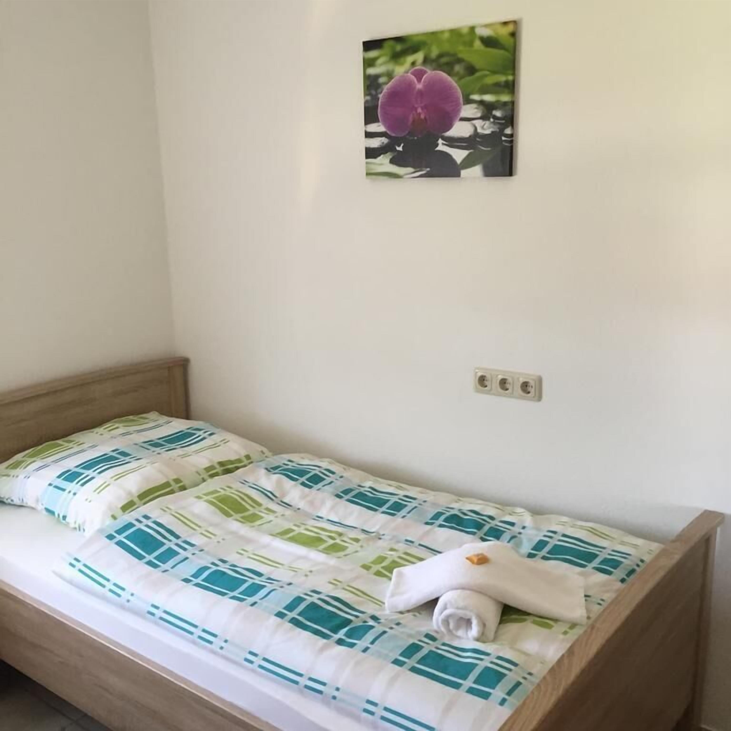 Ein Einzelbett mit einem gemusterten Bettüberwurf, ein aufgerolltes Handtuch und eine Wandsteckdose.