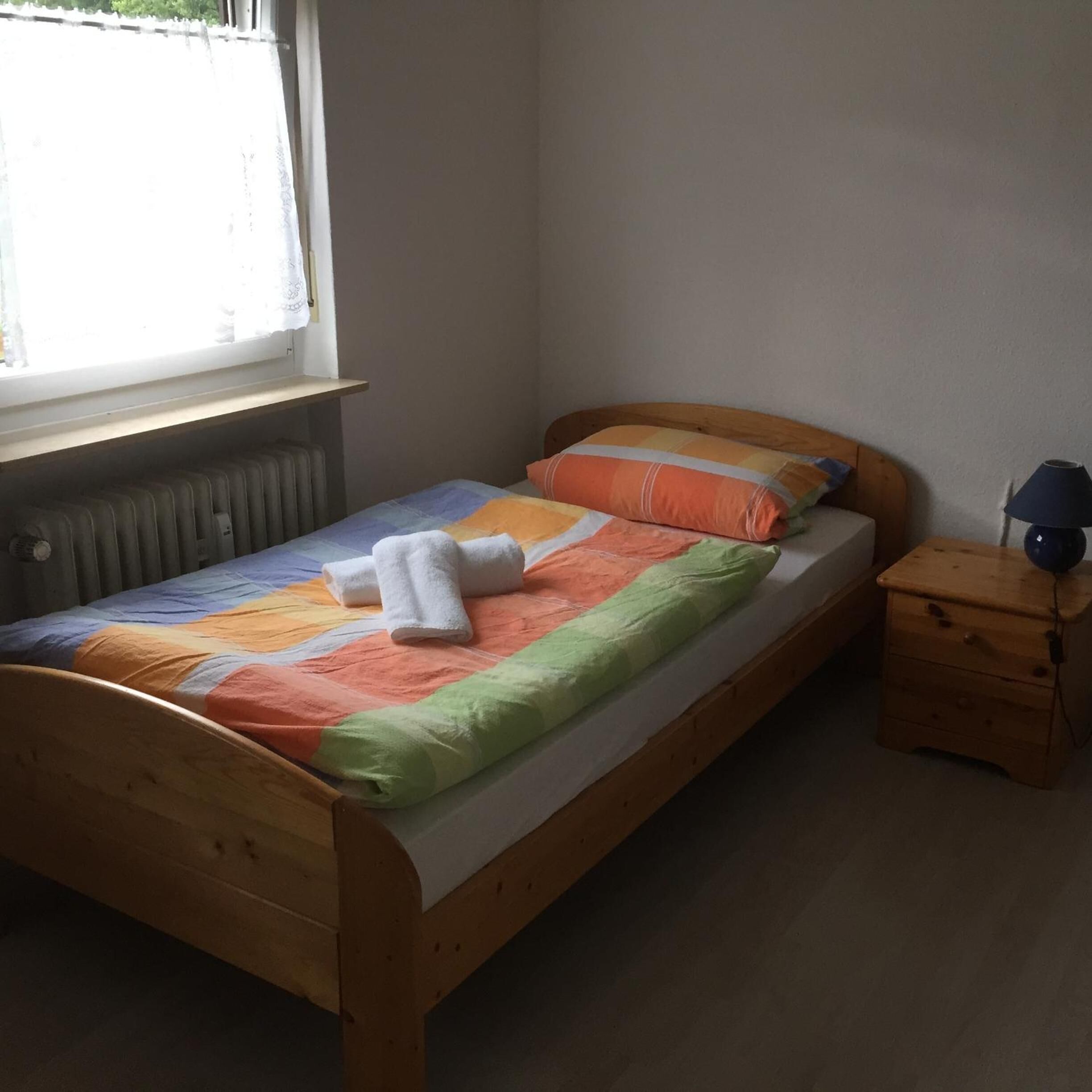 Ein kleines Zimmer mit einem Einzelbett, einem Holzbettgestell, einer bunten Steudecke und einem Nachttisch mit Lampe.