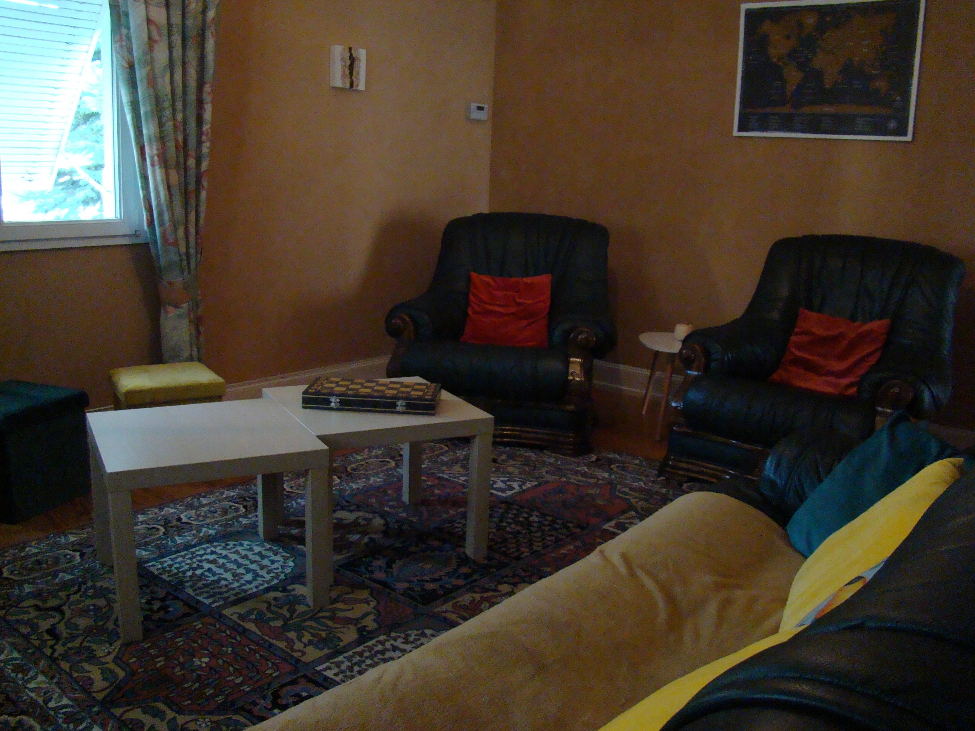 Ein Wohnzimmer mit zwei schwarzen Sesseln, davon einer mit einem roten Kissen, einem beigen Sofa, einem kleinen weißen Tisch und einem gemusterten Teppich.