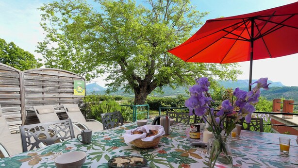 An outdoor dining area with a table set for two, a red umbrella, and a view of the mountains.