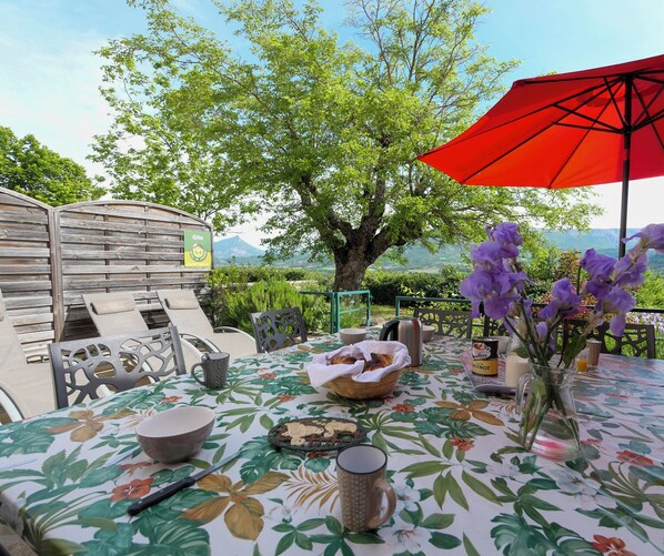 Een terras met een eettafel gedekt voor twee personen, een rode parasol en uitzicht op de bergen.