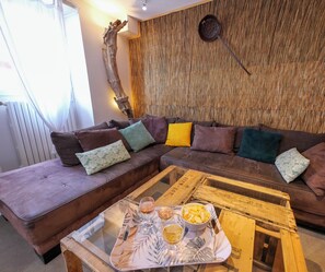 Een gezellige woonkamer met een bruine bank, een houten salontafel met een dienblad vol snacks en drankjes, en een gestructureerde muur achter de bank.