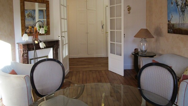 A room with a glass-top dining table, two chairs, a sofa, a small side table with a lamp, a fireplace, a mirror, and a door leading to another room. - Haven of peace in the center of Versailles (Versailles)