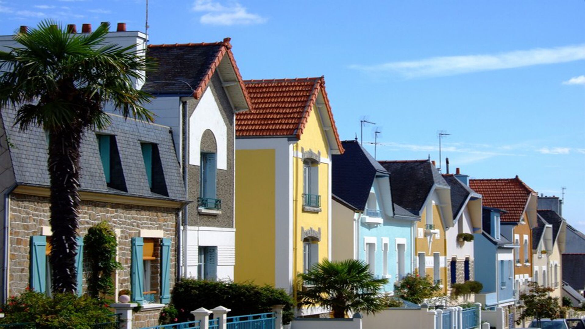 A row of colorful houses with sloped roofs and shutters, a palm tree, and a clear blue sky.
