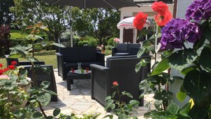 Uma área de pátio externa com mobiliário de vime, piso de ladrilho de pedra e diversos vasos com plantas e flores.
