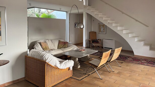 A spacious living area with a high ceiling, a large window, a sofa, a dining table, and a staircase. - Large villa with pool (Saint-Paul-En-Forêt)